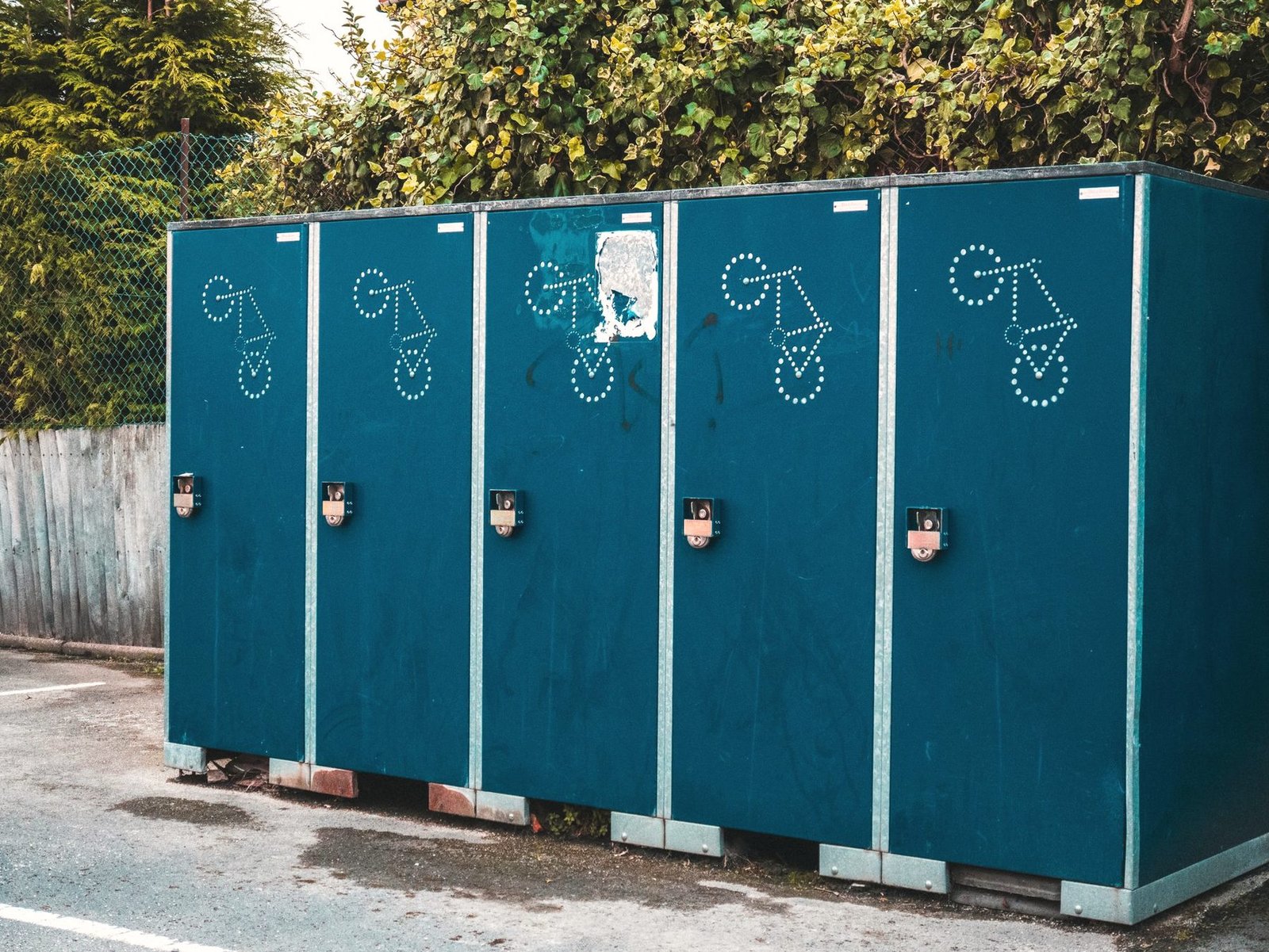 A bank of five teal portable toilet cabins standing against a tall green hedge.