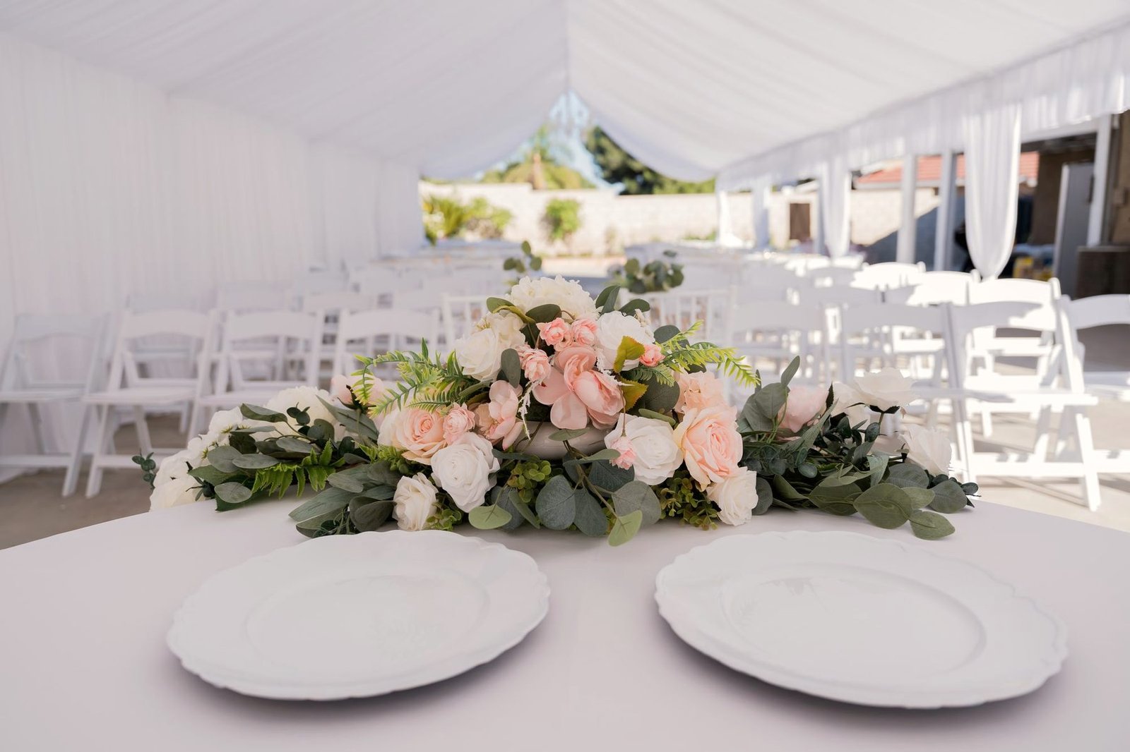 A place setting with white plates and a pale floral arrangement inside a bright marquee.