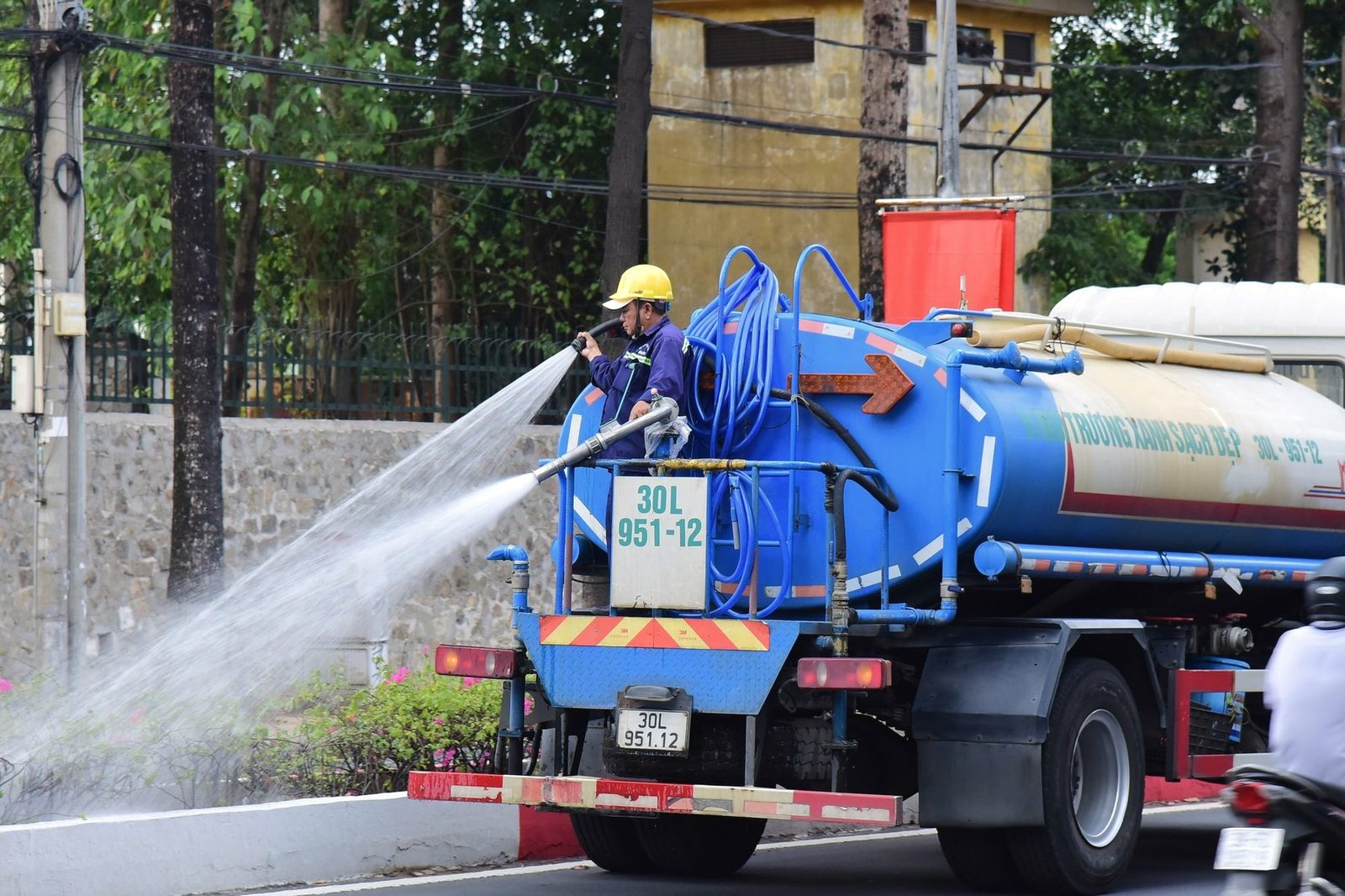 A crew member in a hard hat working a hose from a blue tanker truck on a street.