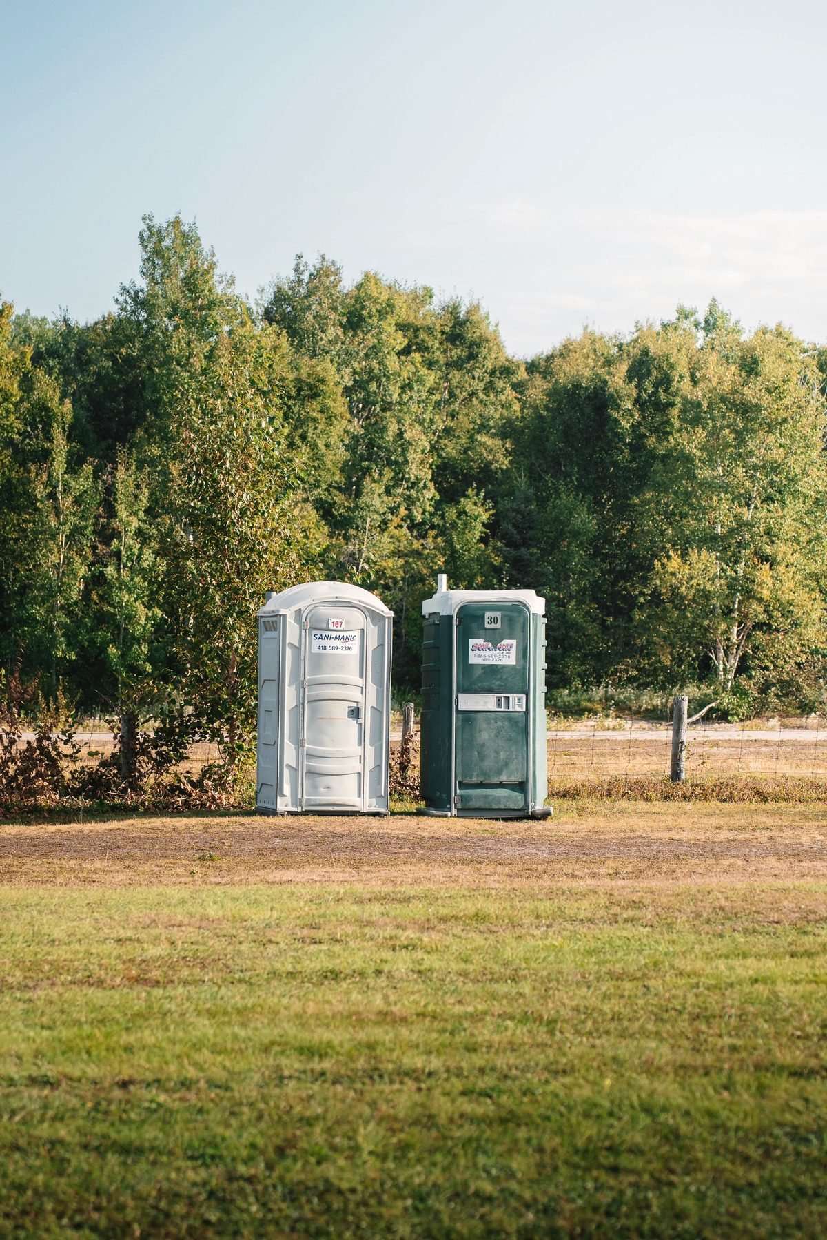 Two portable toilet units standing together on open grass at the edge of woodland.